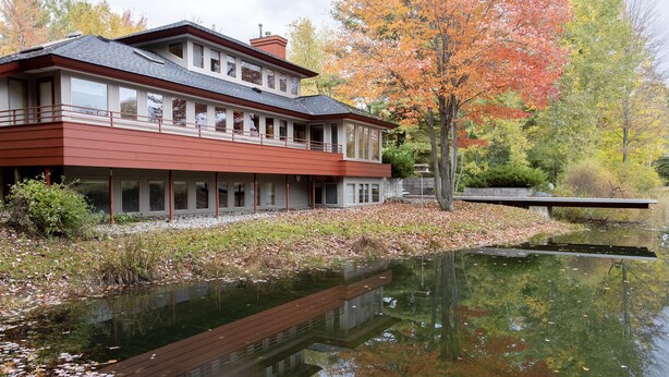 A modern two-story building with a flat roof and large windows, situated next to a calm body of water reflecting the surrounding trees. - Mid-Century Modern Retreat on Private 10 Acres with 1-Acre Pond & Nature Trails. (Midland)