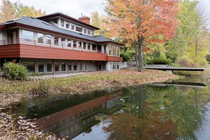 A modern two-story building with a flat roof and large windows, situated next to a calm body of water reflecting the surrounding trees. - Mid-Century Modern Retreat on Private 10 Acres with 1-Acre Pond & Nature Trails. (Midland)