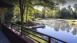 A lakeside deck with a wooden railing, a bench, and a view of a calm lake surrounded by trees. - Mid-Century Modern Retreat on Private 10 Acres with 1-Acre Pond & Nature Trails. (Midland)