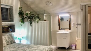 Une chambre à coucher comprenant un lit, une table de chevet, un miroir, une fenêtre avec des stores, un ventilateur de plafond et un radiateur.