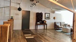 Il s'agit d'une pièce de style « loft », avec un plafond haut, des poutres apparentes et un mur en métal ondulé. On y trouve un banc en bois, une petite table avec une lampe et un canapé avec des coussins.