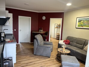 Uma sala de estar moderna com um sofá e uma poltrona cinzentos, uma mesa de centro de madeira e uma pequena mesa lateral. Há uma área de cozinha com fogão e pia, e uma área de jantar com mesa e cadeiras. Um relógio é visível na parede, e um quadro está pendurado acima do sofá.