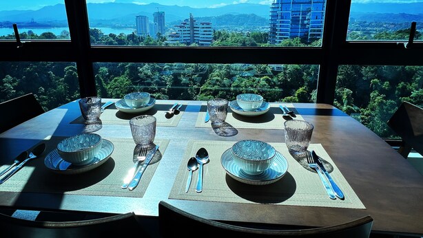 A dining table set with plates, bowls, and cutlery, viewed through large windows overlooking a cityscape and mountains.