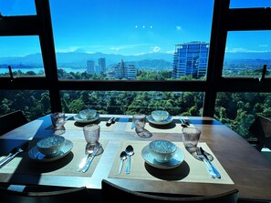 Une table à manger dressée avec des assiettes, des bols et des couverts, vue à travers de grandes fenêtres donnant sur un paysage urbain et des montagnes.