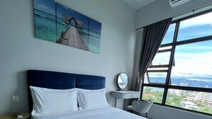 A neatly made bed with white linens and a blue headboard, a small bedside table with a lamp, a round mirror, and a view of the city through large windows.