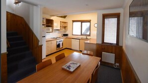 A compact kitchen with a dining area, wooden staircase, and a window with blinds.