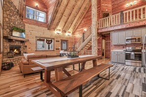 A spacious log cabin interior with a stone fireplace, a wooden dining table, a sofa, and a kitchen area. - Welcome to Heaven in the beautiful Hocking Hills! (Logan)