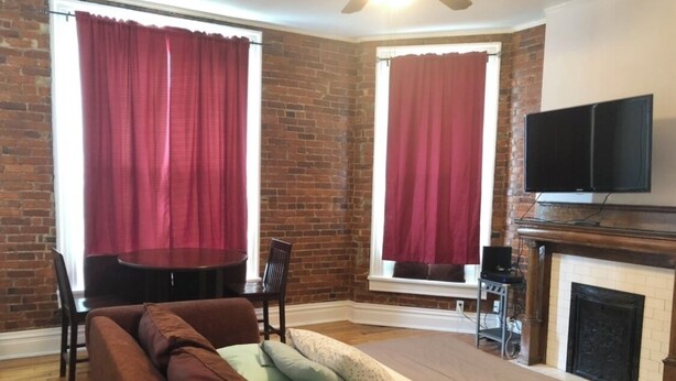 A cozy room with a brick wall, a fireplace, a television mounted above it, and a brown sofa with cushions. - Cantrell’s on Broadway #1 (Nashville)