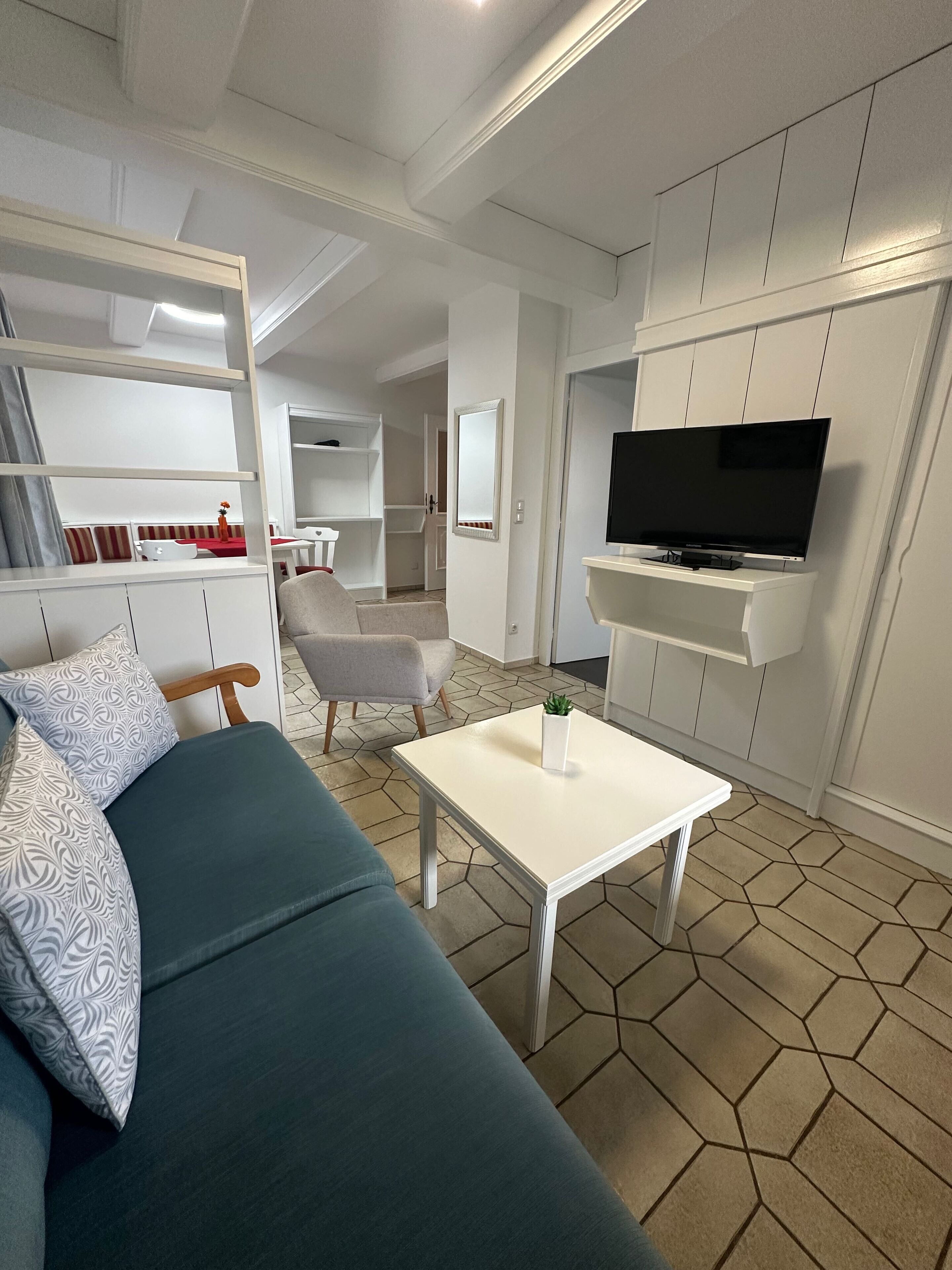 Un salon moderne avec un canapé, une table basse et un téléviseur fixé au mur. On aperçoit en arrière-plan des étagères intégrées et un coin repas.