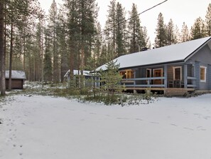 Eine Holzhütte mit einer Holzterrasse, umgeben von verschneiter Landschaft und hohen Kiefern.