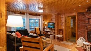 Interior acolhedor de uma cabana de madeira, com sofá, cadeiras, televisão e lareira.