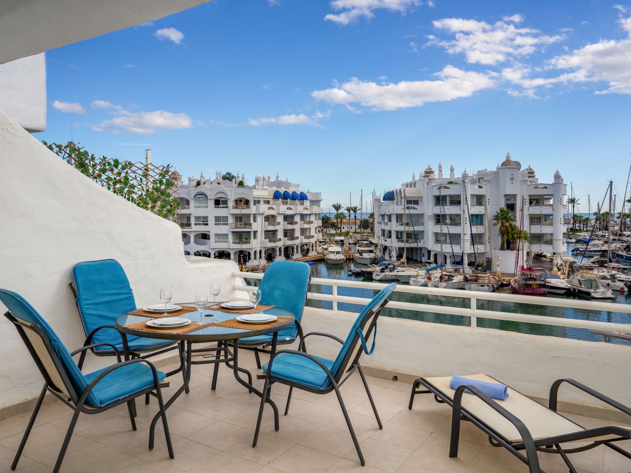 Une terrasse avec une table et des chaises, donnant sur un port de plaisance où sont amarrés des bateaux, et sur un bâtiment blanc doté de balcons.