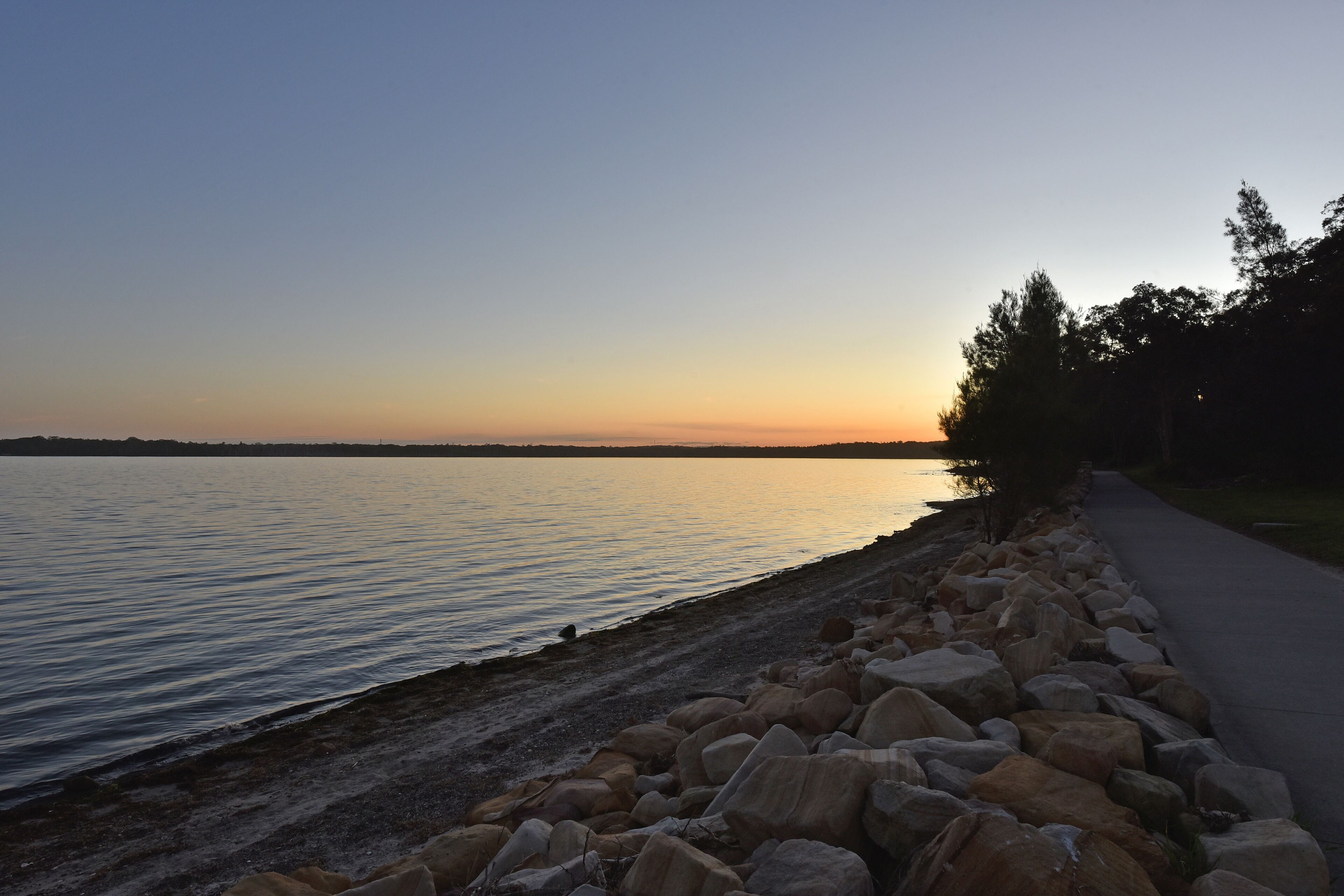 Ein ruhiger See bei Sonnenuntergang mit felsiger Uferlinie und einem befestigten Weg.