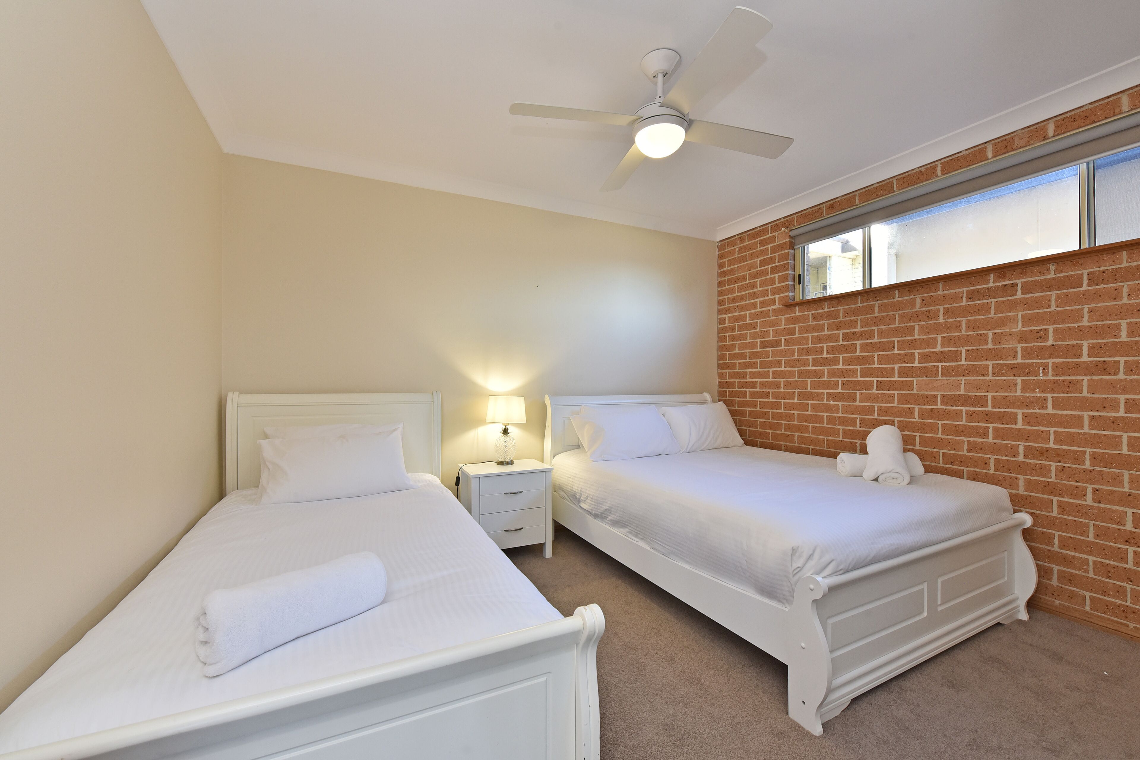 A bedroom with two single beds, a ceiling fan, a nightstand with a lamp, and a brick wall.