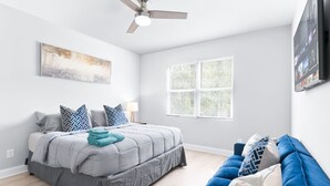 A bedroom with a bed, a blue couch, a ceiling fan, a window, and a flat-screen TV.