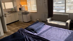 A compact hotel room with a kitchenette, a bed with a blue blanket, a white armchair, and a window with blinds. - Zenibako House 3F-RoomA (Otaru)