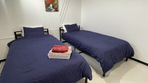 Dos camas individuales con ropa de cama azul oscuro, una pared blanca con un cuadro y una pequeña bandeja blanca con toallas.