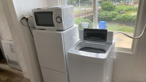 Een kleine keuken met een magnetron bovenop een koelkast, een wasmachine en een raam met uitzicht op groen en een treinspoor.
