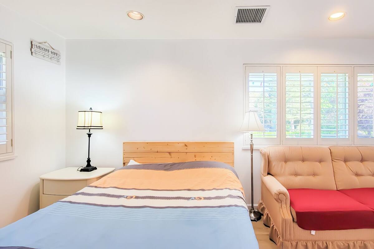 A bedroom with a bed, a chair, a nightstand, a lamp, and a window with blinds.