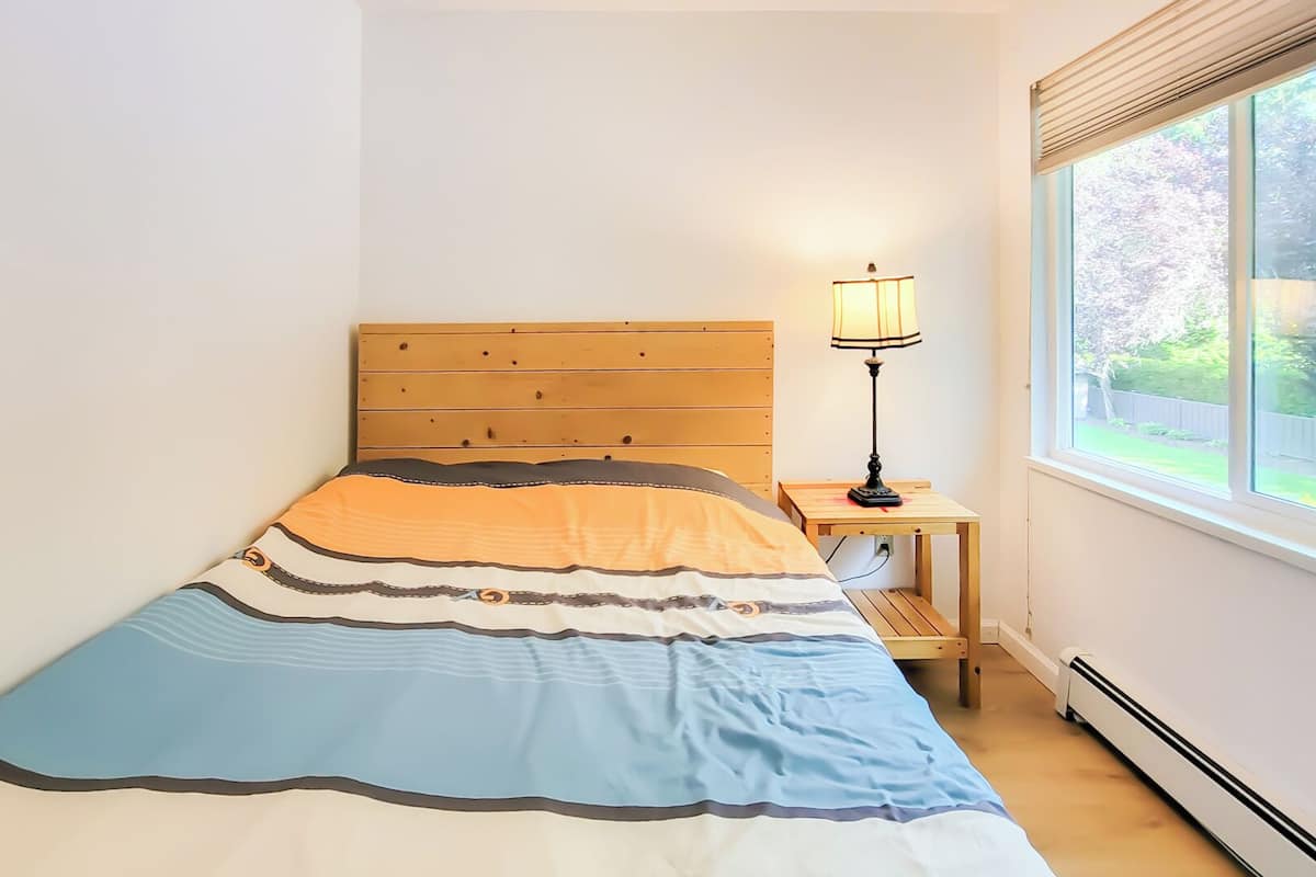 A bedroom with a wooden bed, a bedside table with a lamp, and a window with a view of greenery.