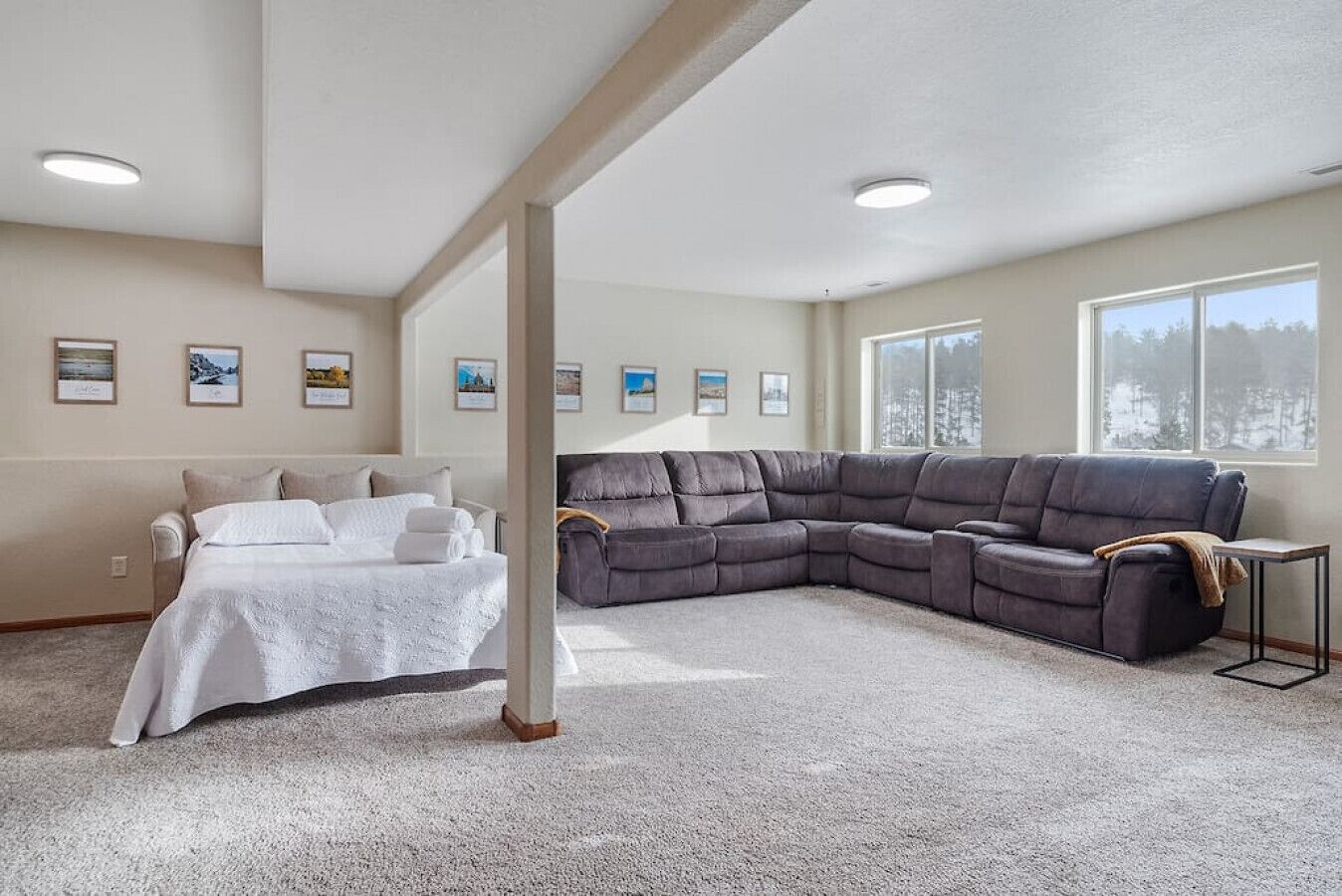 A spacious living room with a sectional sofa, a bed with a white bedspread, and framed pictures on the walls.