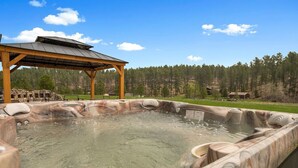 Un espace avec un bain à remous, surmonté d’une pergola en bois, et entouré d’un paysage naturel avec des arbres et un ciel dégagé.
