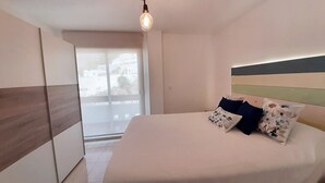 Une chambre à coucher avec un lit, une table de chevet, une fenêtre donnant sur des immeubles et un luminaire suspendu.