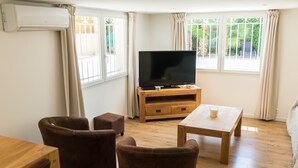 Een woonkamer met een flatscreen televisie op een houten standaard, een salontafel en twee bruine leren fauteuils.