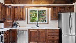 A kitchen with wooden cabinets, a stainless steel refrigerator, and a window with a view of trees.