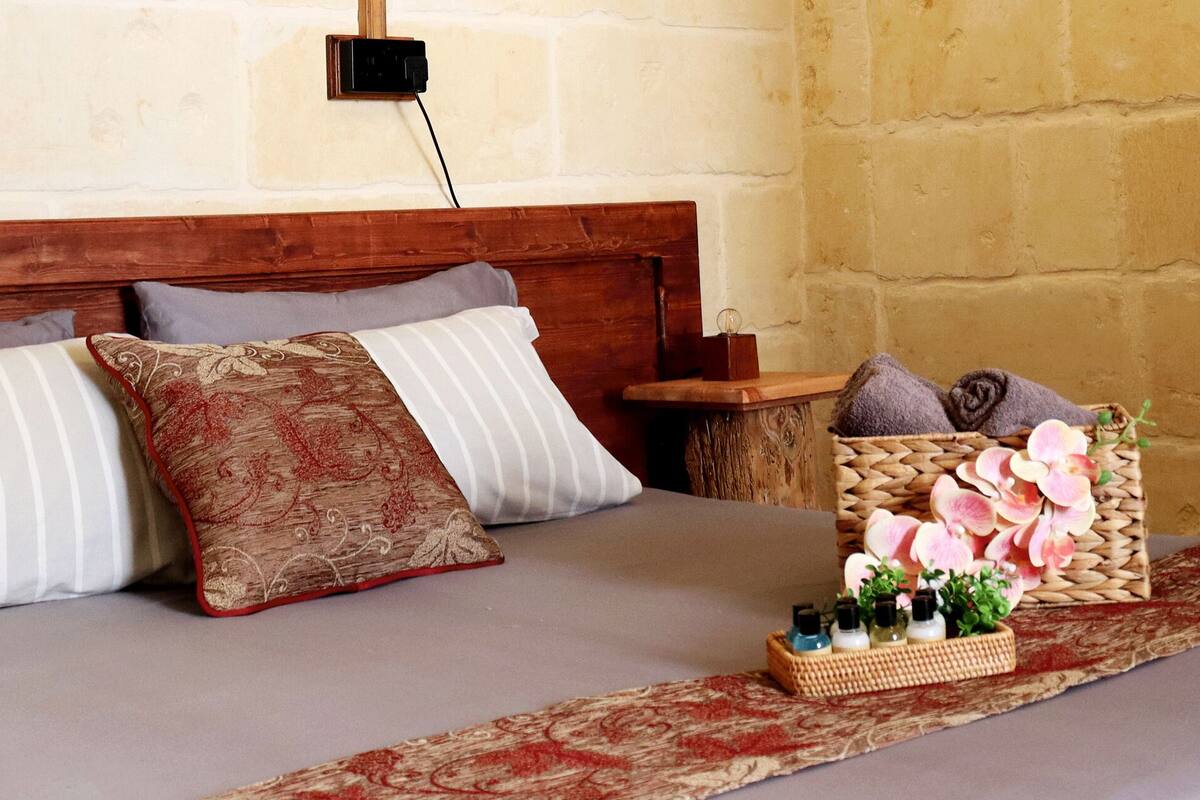 Una cama bien hecha con almohadas a rayas y estampadas, una cesta tejida con flores y artículos de aseo, y una mesita de noche de madera con una lámpara.