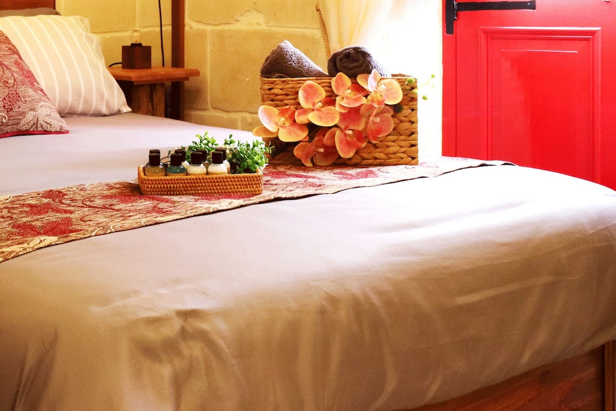Una cama bien hecha con un cojín decorativo, una mesita de noche pequeña y un cesto de artículos de aseo.