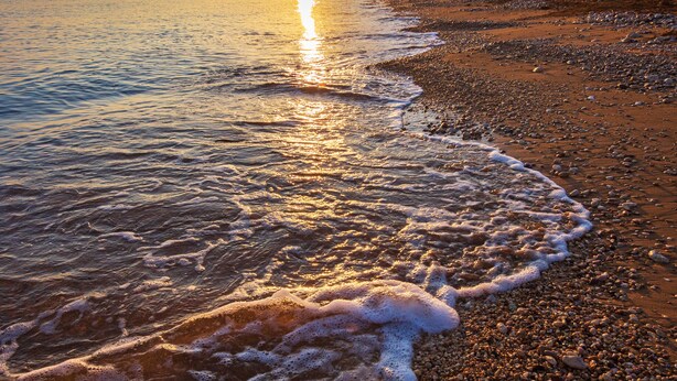 Una playa pedregosa con el sol poniéndose en el horizonte, mar tranquilo y un acantilado distante.