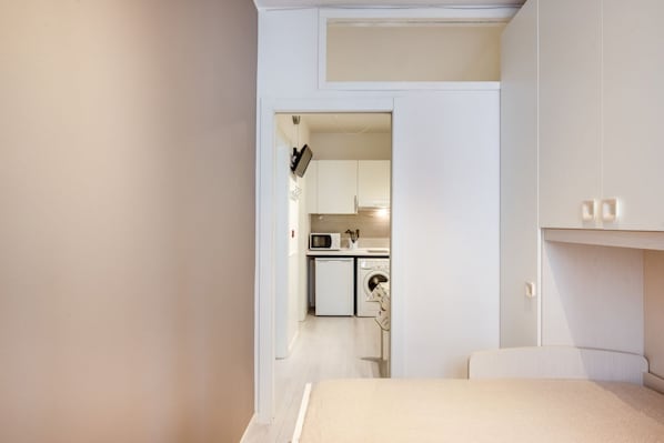 Un dormitorio con cama, armario y vista a una cocina con electrodomésticos.