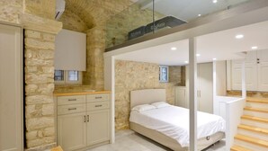 Un dormitorio con pared de piedra, una cama, una escalera de madera y un balcón con cerramiento de vidrio.