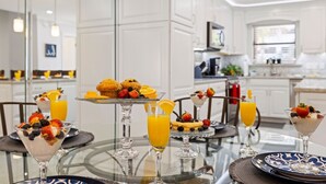 A modern kitchen with white cabinetry, a glass dining table, and various breakfast items including muffins, fruit, and beverages. - MerSea Beaucoup @ Gulf Highlands Sleeps 6 (Panama City Beach)