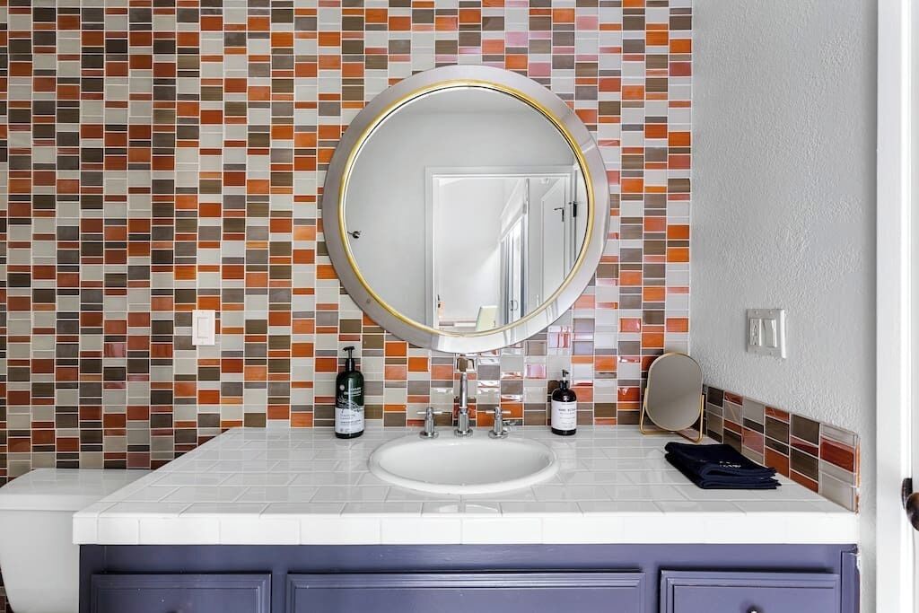 Une salle de bain avec un mur recouvert de carreaux, un miroir rond, un lavabo blanc et un meuble-lavabo avec des tiroirs.