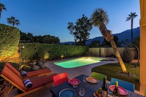 A poolside area with lounge chairs, a dining table with plates and glasses, and a well-maintained garden with a hedge and palm trees.