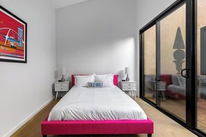 A bedroom with a bed, two bedside tables, a nightstand, a large window with a view, and a framed picture on the wall.