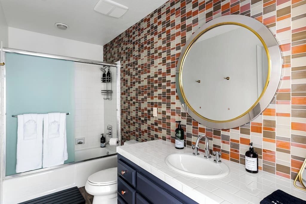 A bathroom with a tiled mosaic wall, a round mirror with a gold frame, a white sink, and a glass shower enclosure.