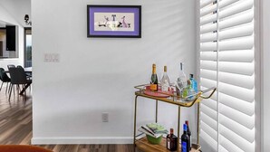 A room with a white wall, a framed picture, a cart with bottles, and a window with blinds. - Stylish Palm Springs Retreat with Pool (Palm Springs)