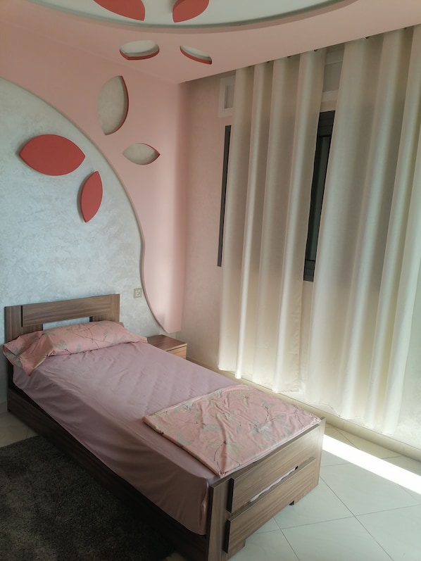 A bedroom with a bed, a nightstand, a wooden dresser, and a window with curtains.