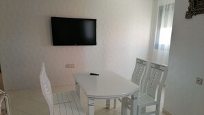 A dining area with a white table and chairs, a flat-screen TV mounted on the wall, and a window with sheer curtains. - Boujemaa's house 2 (Tanger)