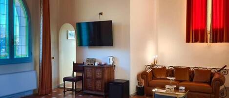 A living room with a large window, a flat-screen TV mounted on the wall, a brown leather sofa, a wooden cabinet, a small table, and a patterned rug.