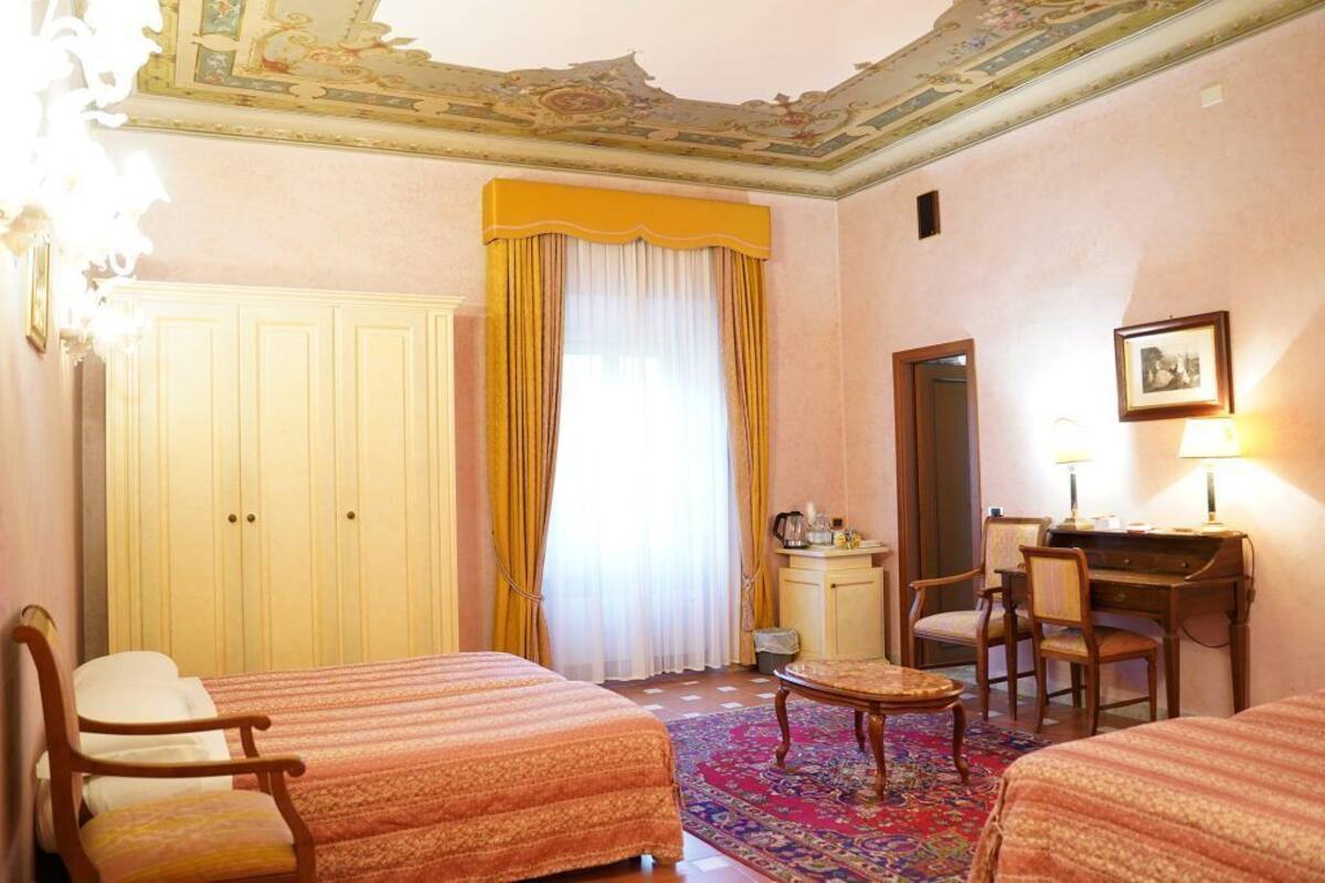 Ein Hotelzimmer mit zwei Betten, einem Schreibtisch, einem Stuhl, einem kleinen Tisch, einem Kleiderschrank und einem Fenster mit Vorhängen.
