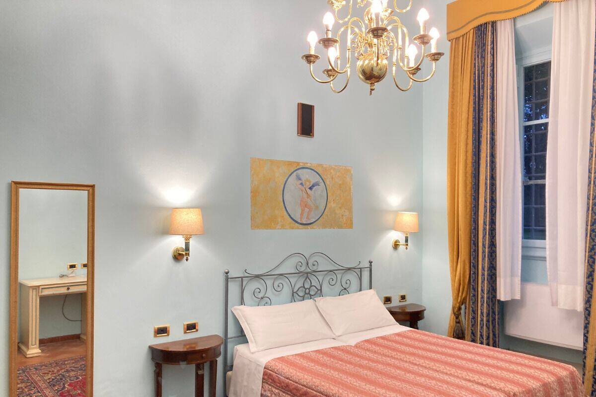 Ein Schlafzimmer mit Bett, zwei Nachttischlampen, einer Chandelieber, einem Spiegel und einem Fenster mit Vorhängen.