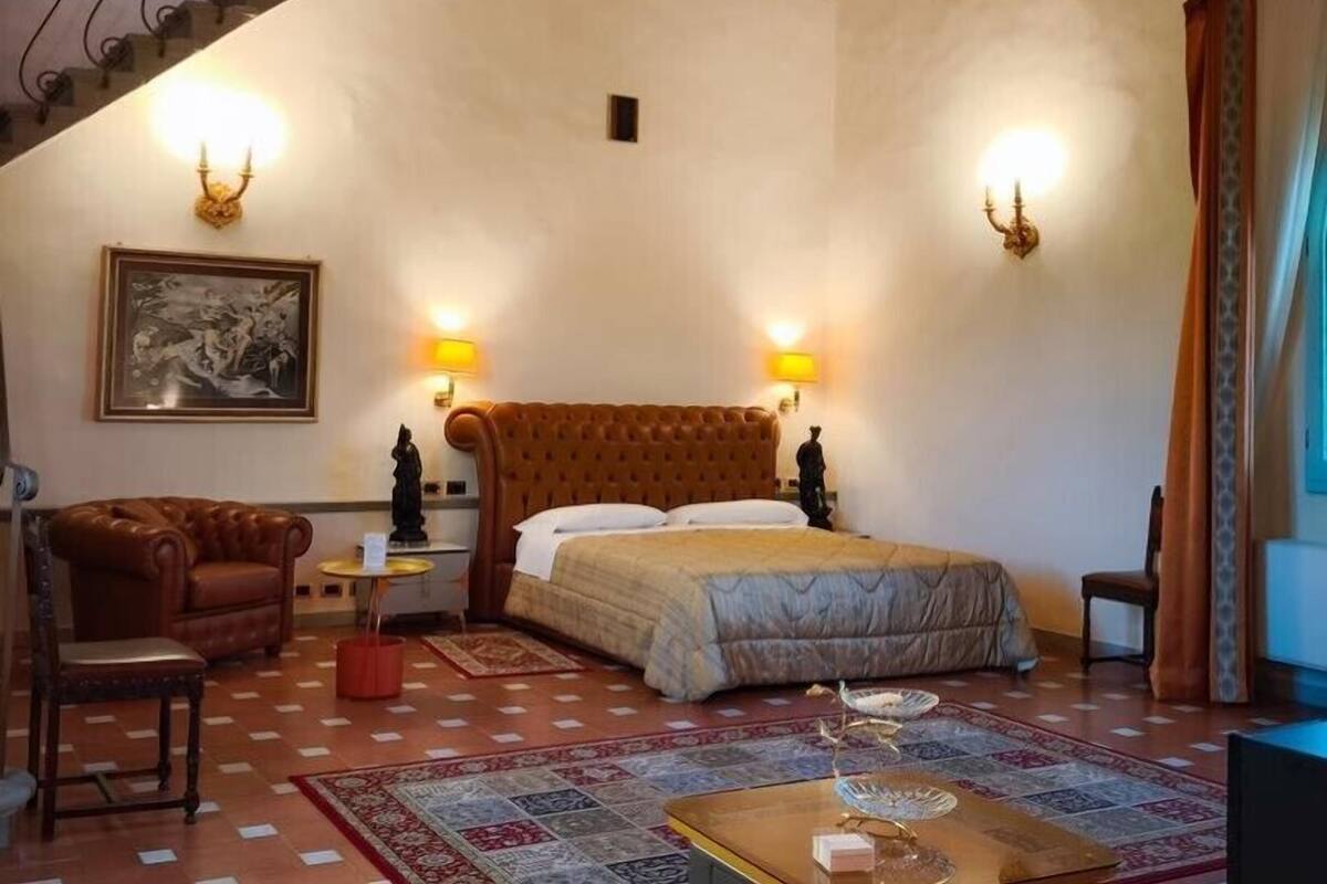 Ein Schlafzimmer mit einem großen Bett, zwei Wandlampen, einem Gemälde, einem Sessel, einem kleinen Tisch und einem gemusterten Teppich.