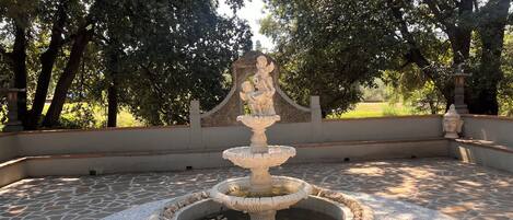 Una fuente de piedra con una estatua en el centro, rodeada por un borde circular de piedra y guijarros.
