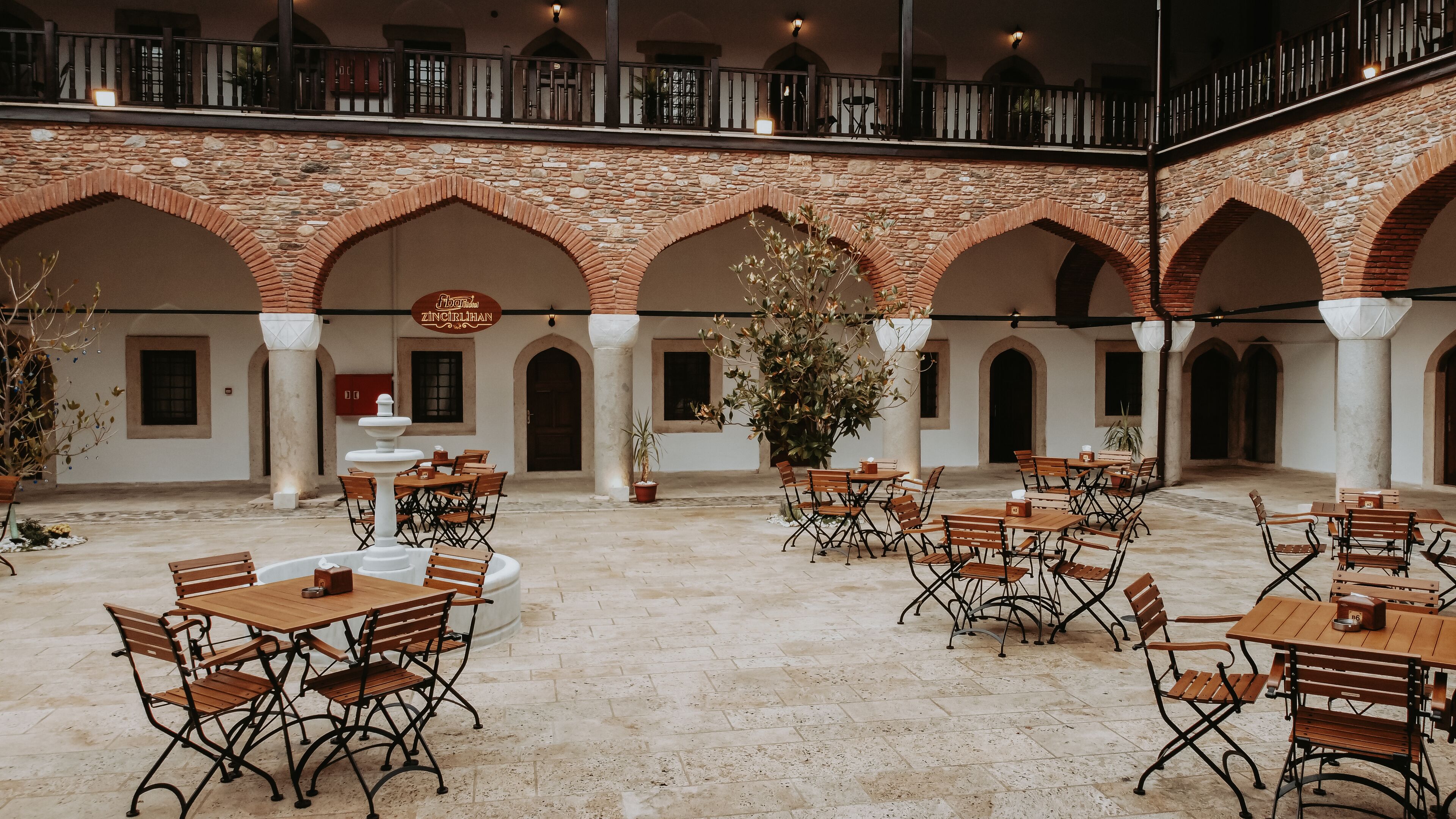 A spacious courtyard with stone arches, multiple wooden tables and chairs, and a central fountain.