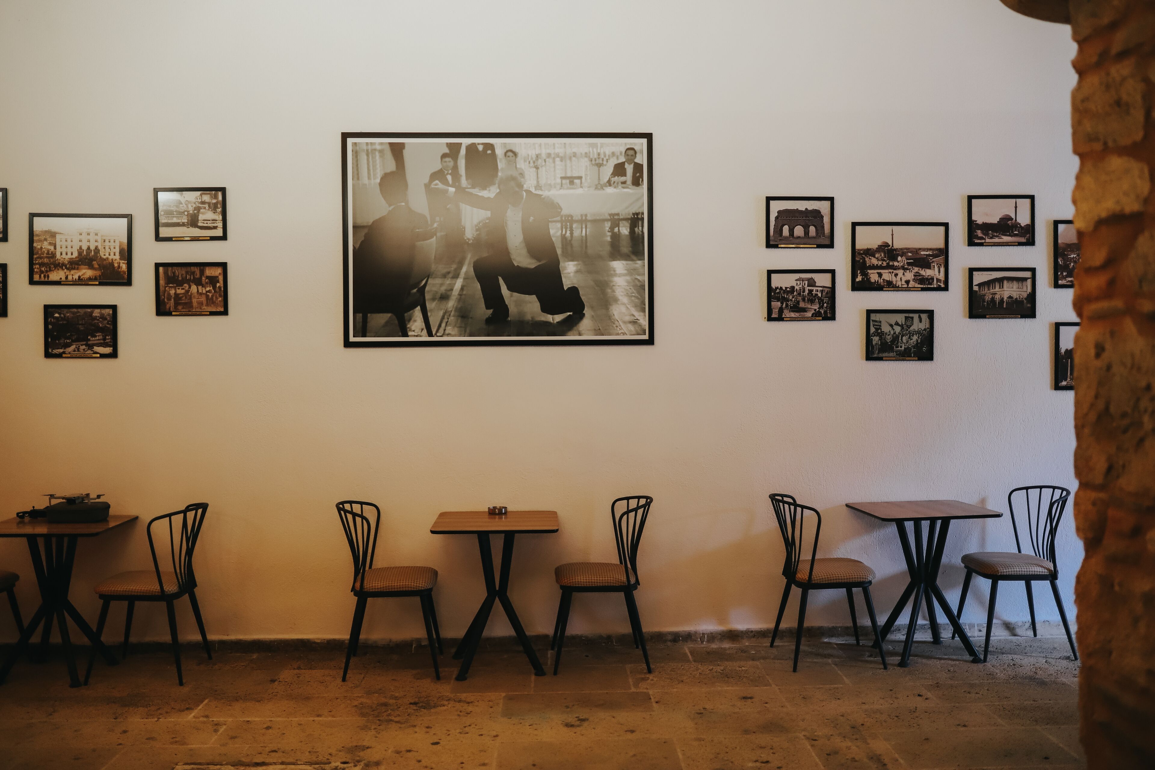 A café with a rustic stone floor, black metal chairs, and tables. There are framed photographs on the wall.