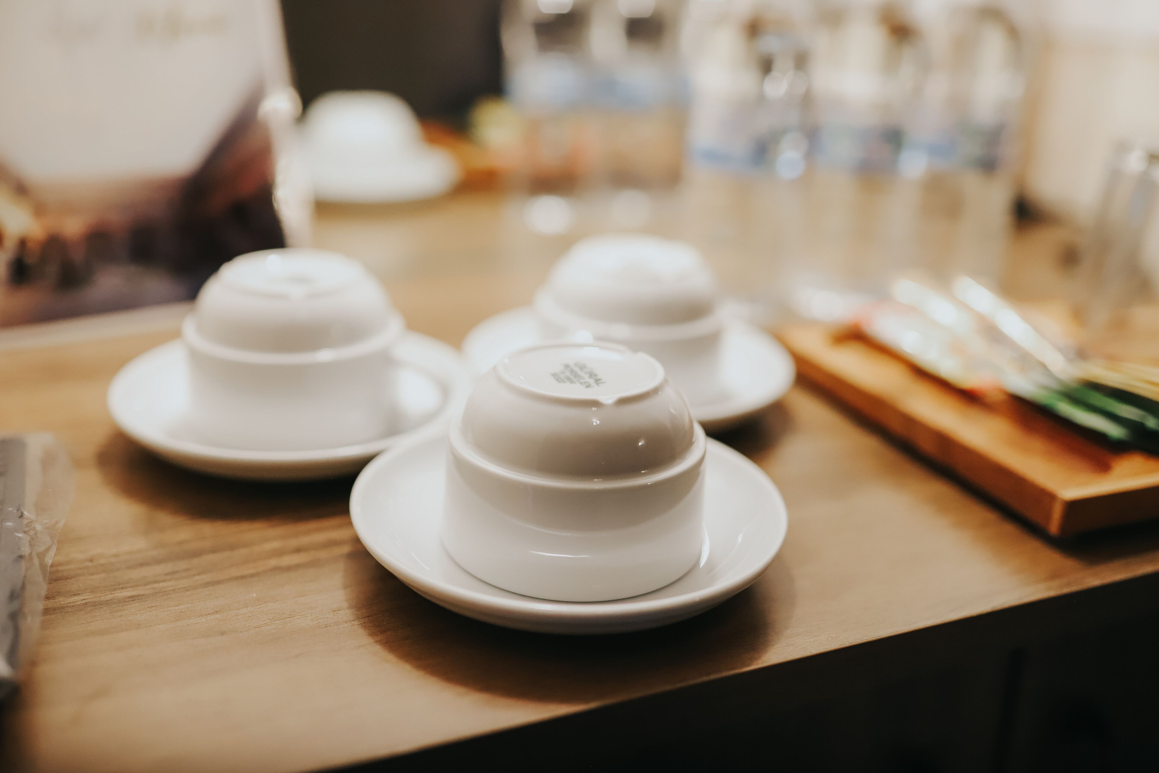 A table with four white teapots on saucers, a wooden cutting board with a knife, and a bottle of water in the background.